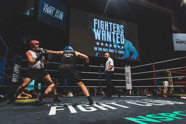 Two boxers in head guards exchange punches, with the White Collar Fight Club screen behind them