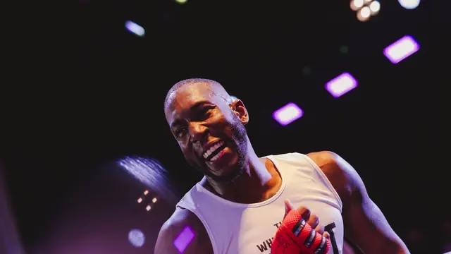 A fighter in a white vest points across the ring under purple lights