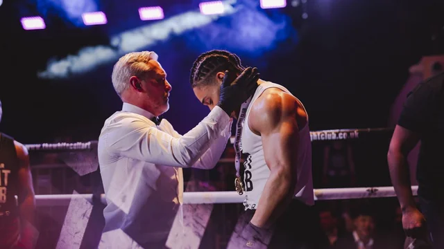 A referee in a white shirt checks a fighter's head guard in the ring