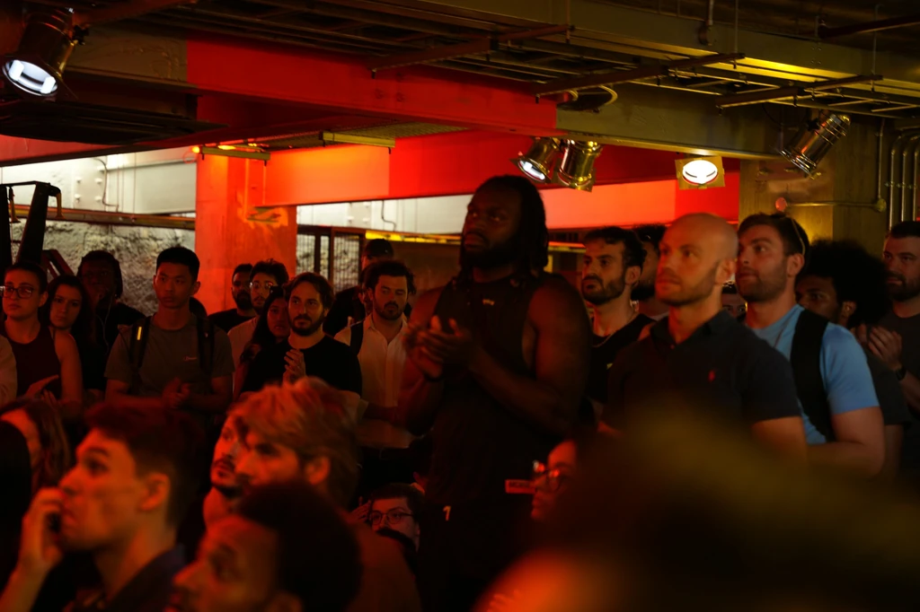 A packed room watching intently under red lights