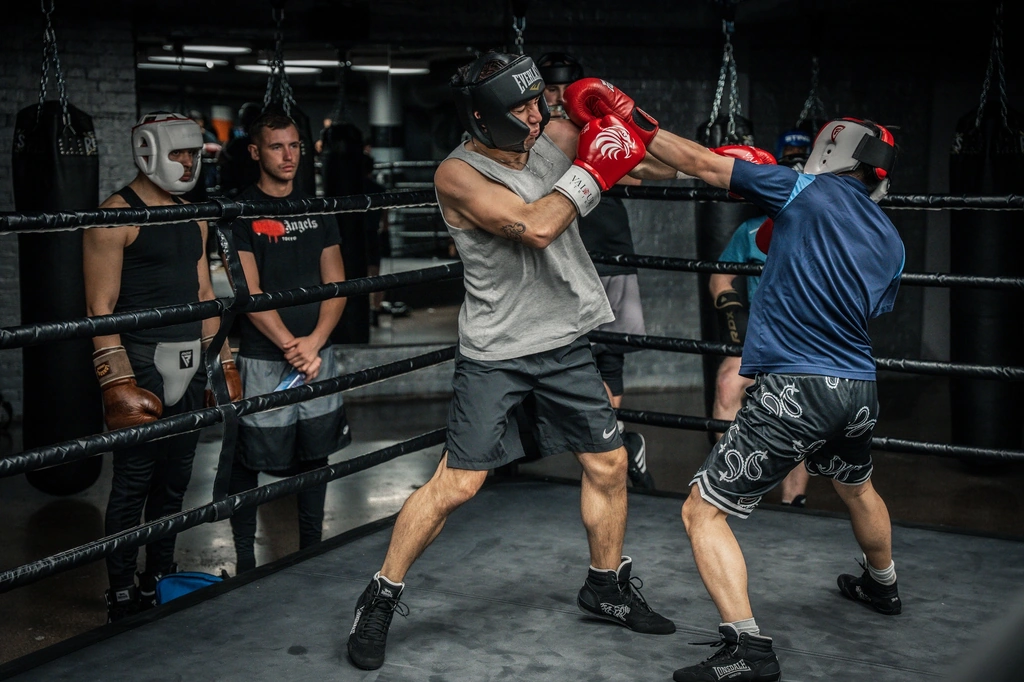 Two men in head guards sparring in the ring while others wait on the ropes