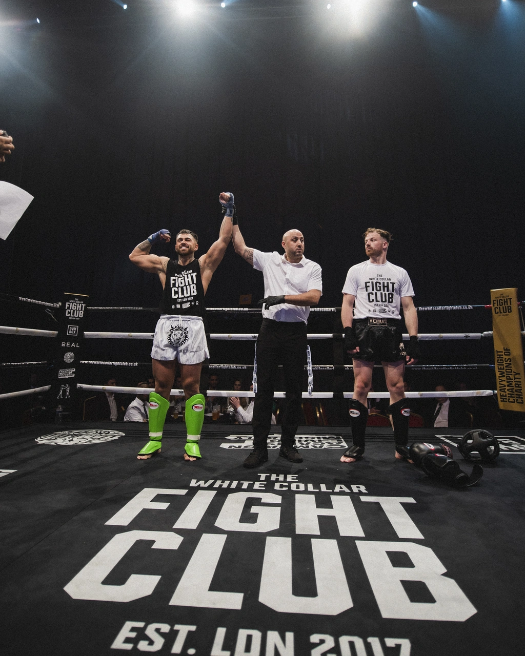 A winner's hand raised by the referee, standing on the White Collar Fight Club branded ring canvas